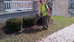 A person wearing a yellow vest and red cap uses a leaf blower near a brick building and neatly trimmed bushes.