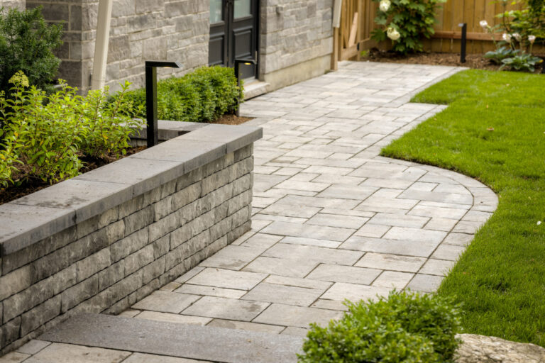 Stone pathway alongside a modern building, bordered by greenery and a stone wall, leading towards a wooden fence in a landscaped garden setting.