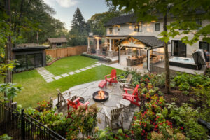 A backyard features a patio with chairs around a fire pit, lush garden, modern house, large deck, and surrounding trees. No landmarks visible.