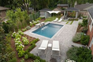 Backyard with a small rectangular pool surrounded by greenery, lounge chairs, and a patio. A modern garden features flowers and trees.